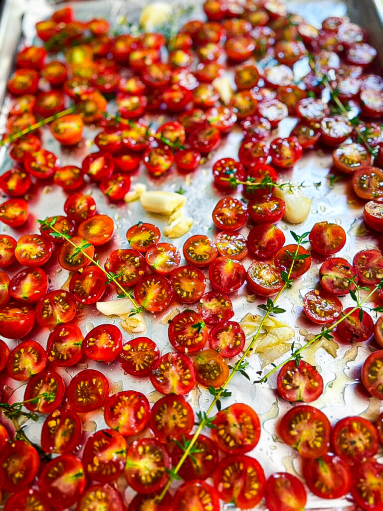 A baking tray filled with halved cherry tomatoes, garlic cloves, and sprigs of thyme, ready for roasting.