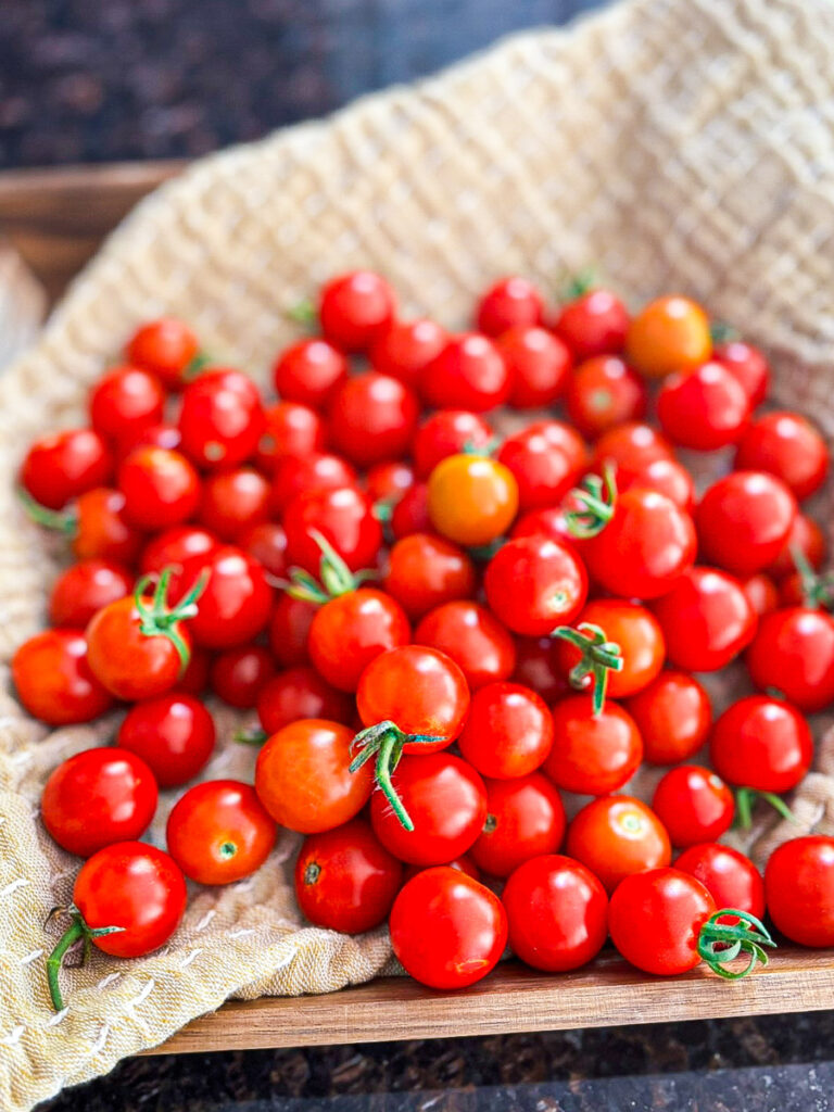 A close-up image of a wooden tray filled with an abundant harvest of ripe cherry tomatoes, showcasing various shades of red and a few orange ones, resting on a textured beige cloth.