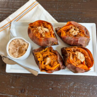 Four baked sweet potatoes topped with creamy harvest honey butter, served on a white platter with extra whipped honey butter on the side.