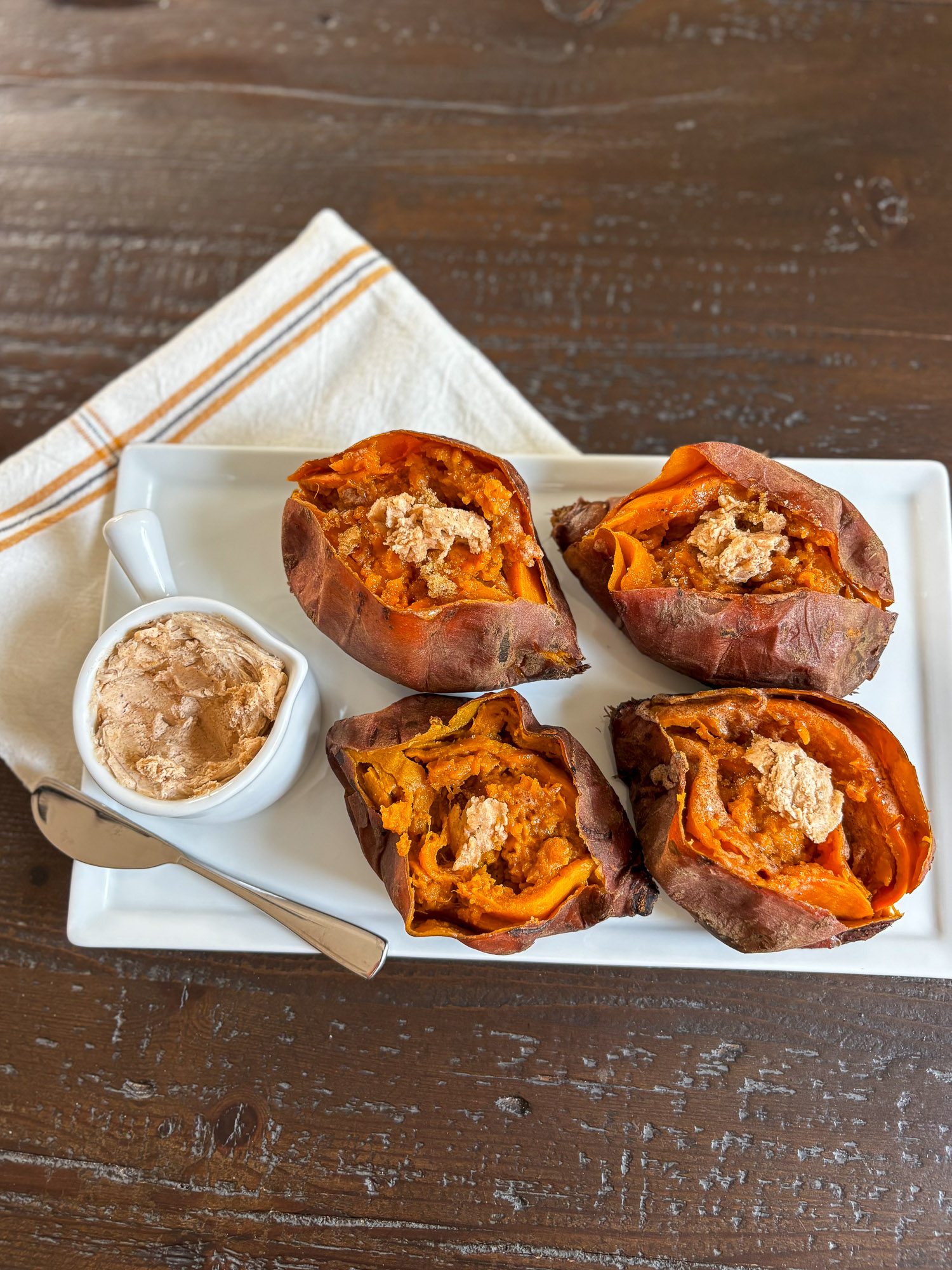 Four baked sweet potatoes topped with creamy harvest honey butter, served on a white platter with extra whipped honey butter on the side.