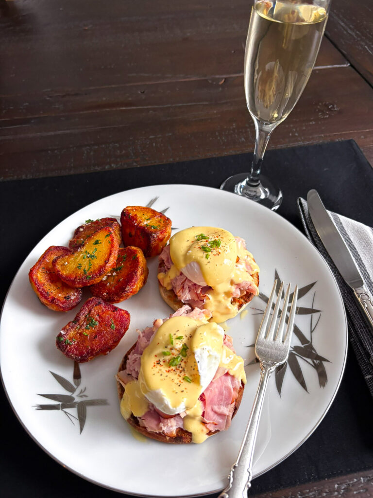 A plated serving of Eggs Benedict featuring poached eggs, Canadian bacon on toasted English muffins, topped with hollandaise sauce, accompanied by crispy fried potatoes and a glass of champagne.