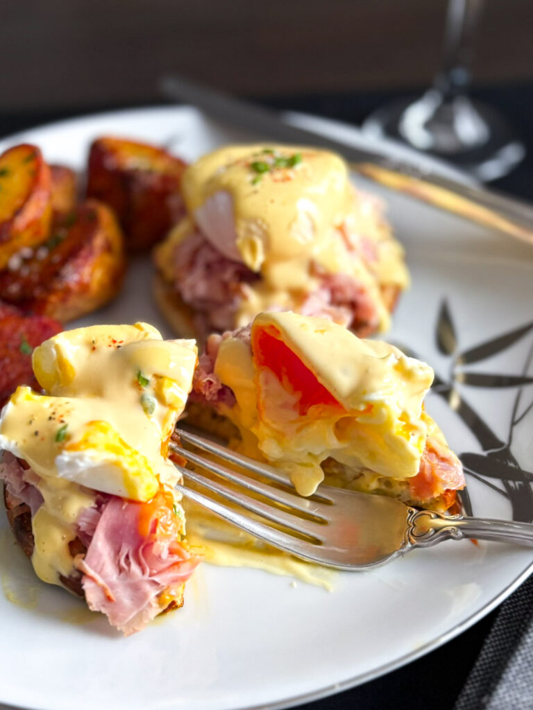 A close-up view of a plated Eggs Benedict with poached eggs, Canadian bacon, and creamy hollandaise sauce, served alongside roasted potatoes.