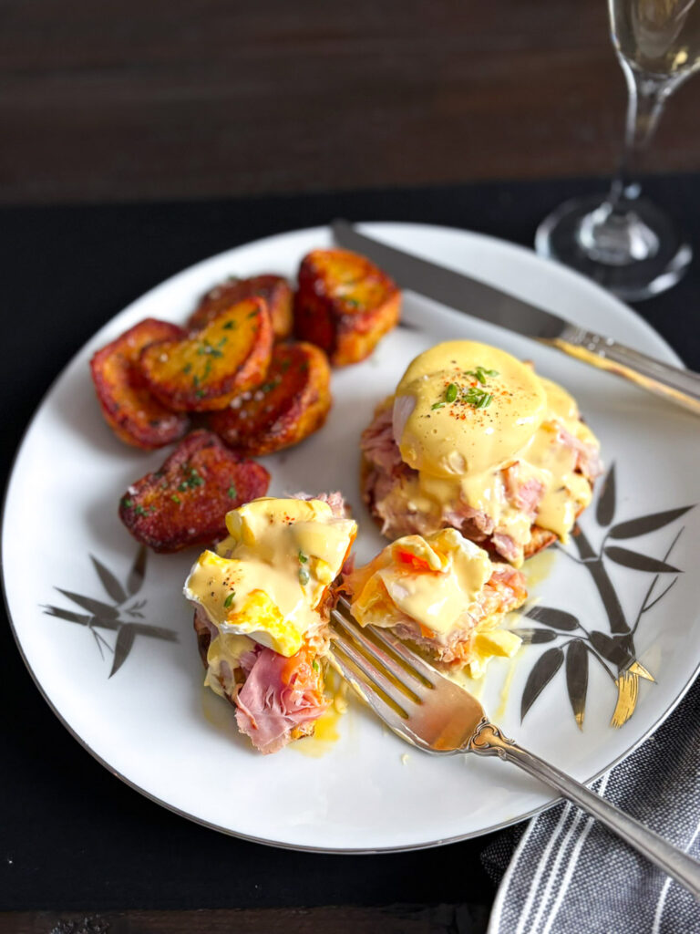 A close-up of a plate featuring Eggs Benedict topped with hollandaise sauce, served with slices of ham and golden-brown roasted potatoes on the side.