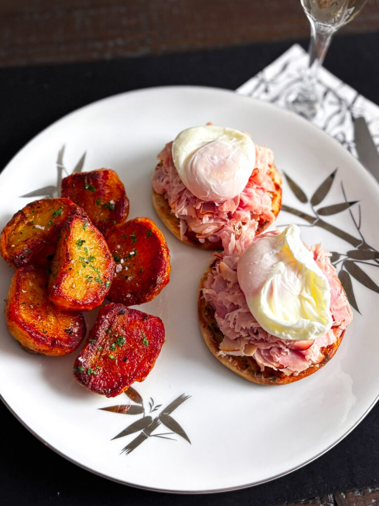 A beautifully plated Eggs Benedict dish featuring poached eggs on toasted English muffins topped with shredded ham, accompanied by crispy roasted potatoes.