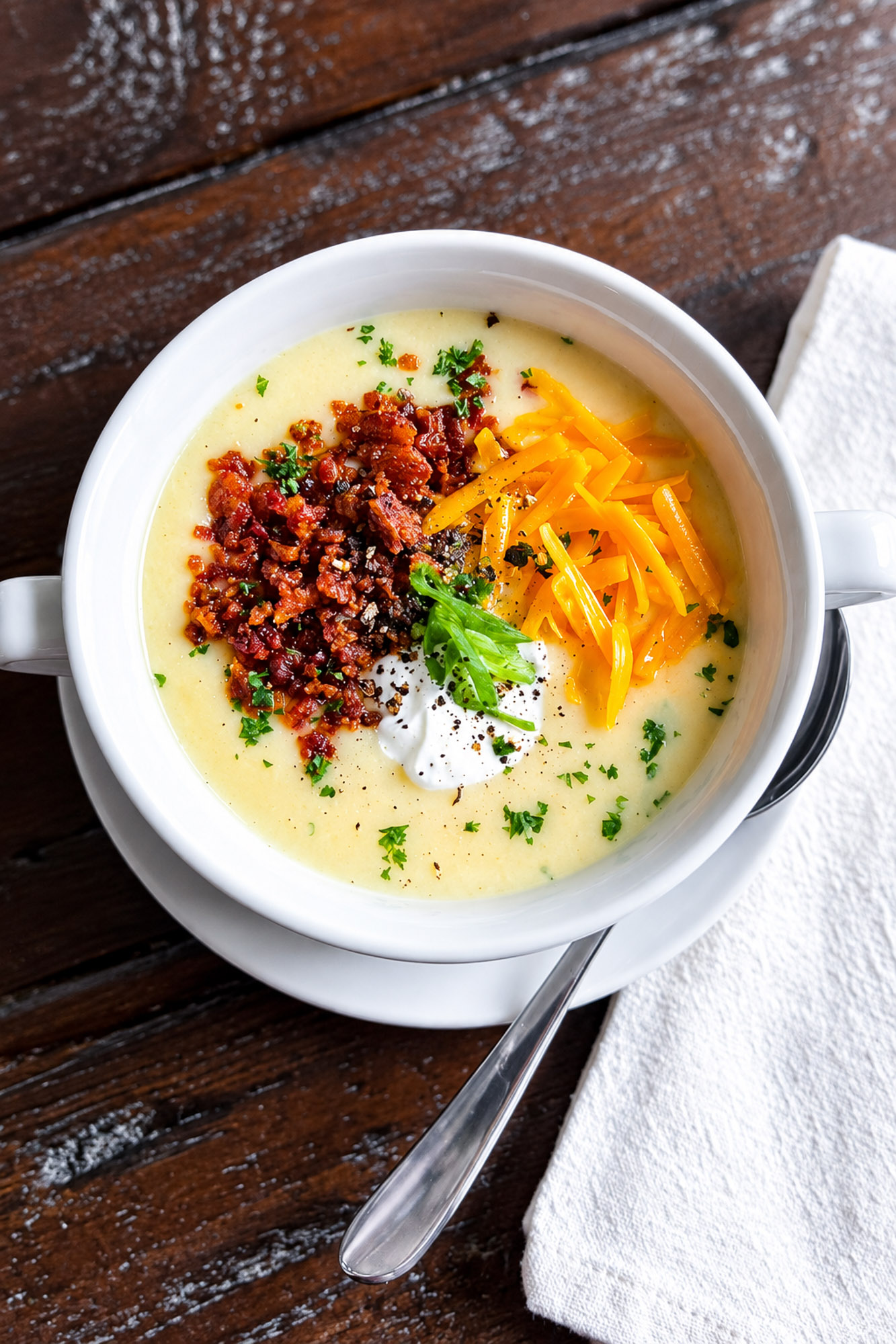 Creamy potato soup topped with bacon, shredded cheddar, sour cream, green onions, parsley, and black pepper in a white bowl.