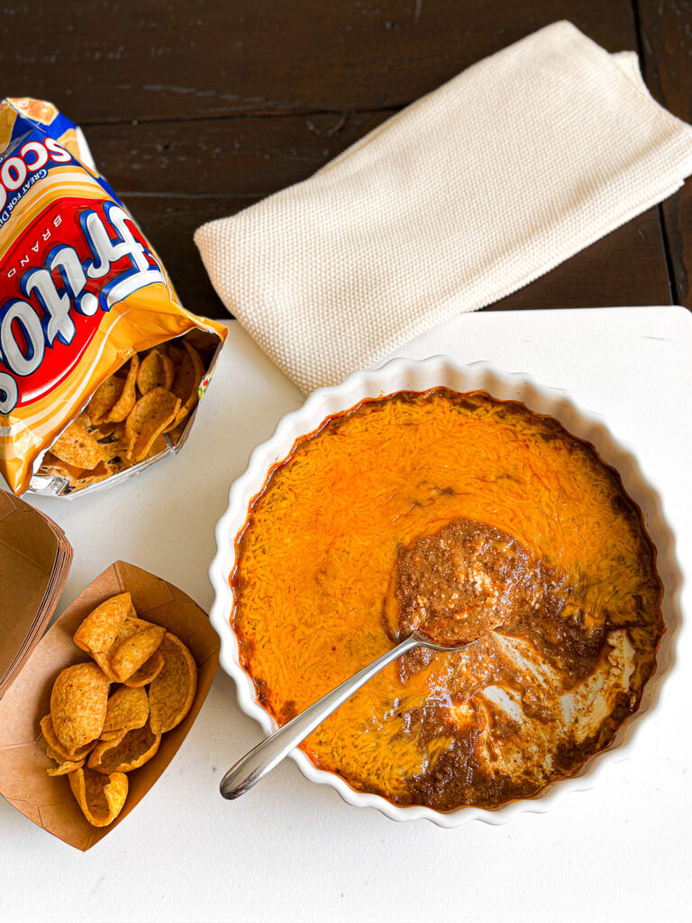 Classic Cincinnati chili dip topped with melted cheddar, served with Fritos Scoops and a spoonful lifted from the dish.