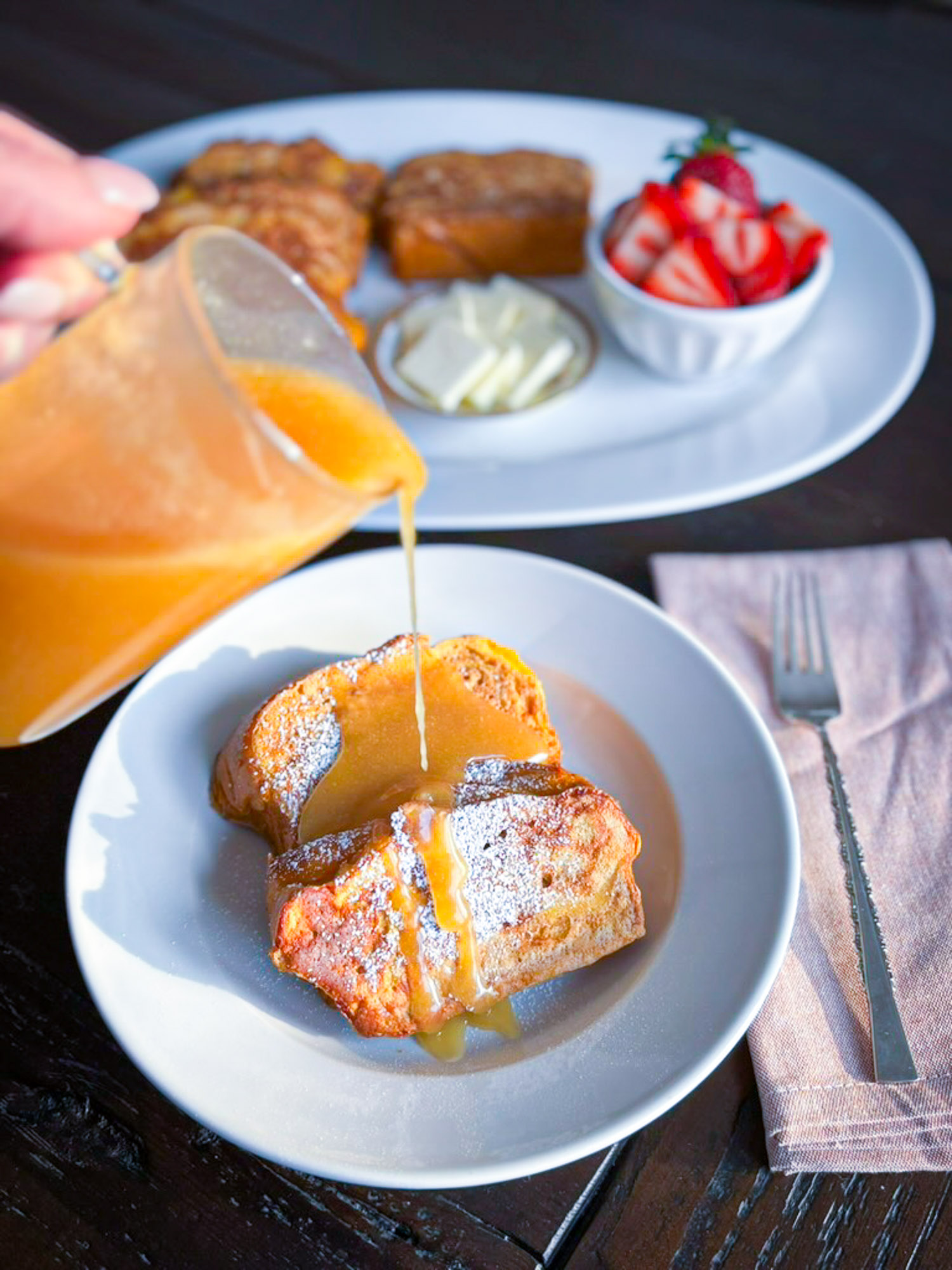 A plate of French toast topped with powdered sugar and drizzled with syrup, accompanied by a bowl of strawberries, butter, and additional French toast slices in the background.