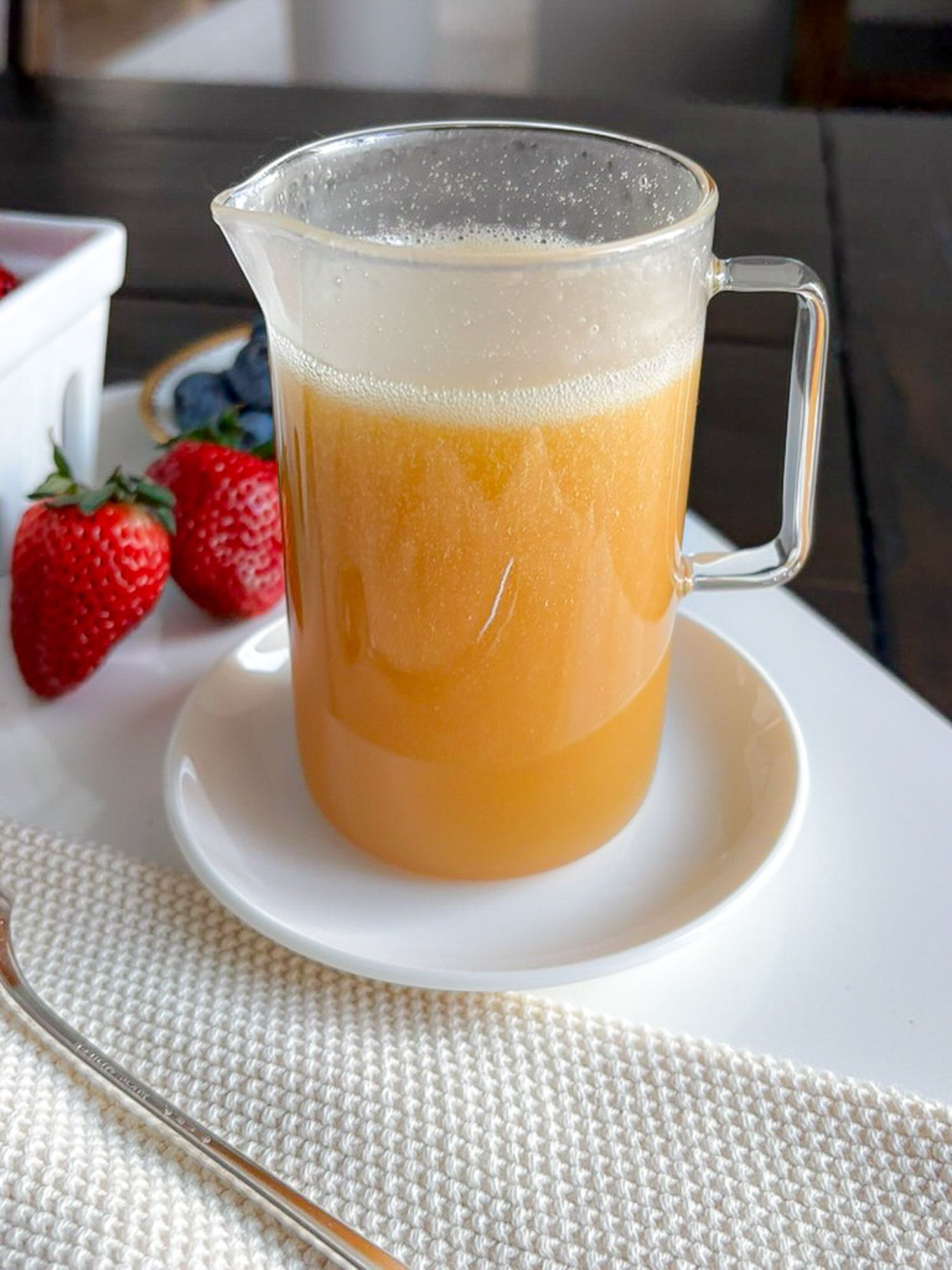 A clear glass jug filled with light brown liquid, sitting on a small white plate. Fresh strawberries are placed on the side, with a spoon resting on a textured white placemat in the foreground.