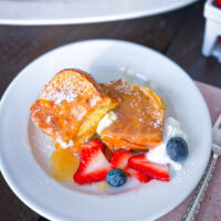 Thick slice of French toast drizzled with buttermilk syrup on a white plate with strawberries, blueberries and cream.