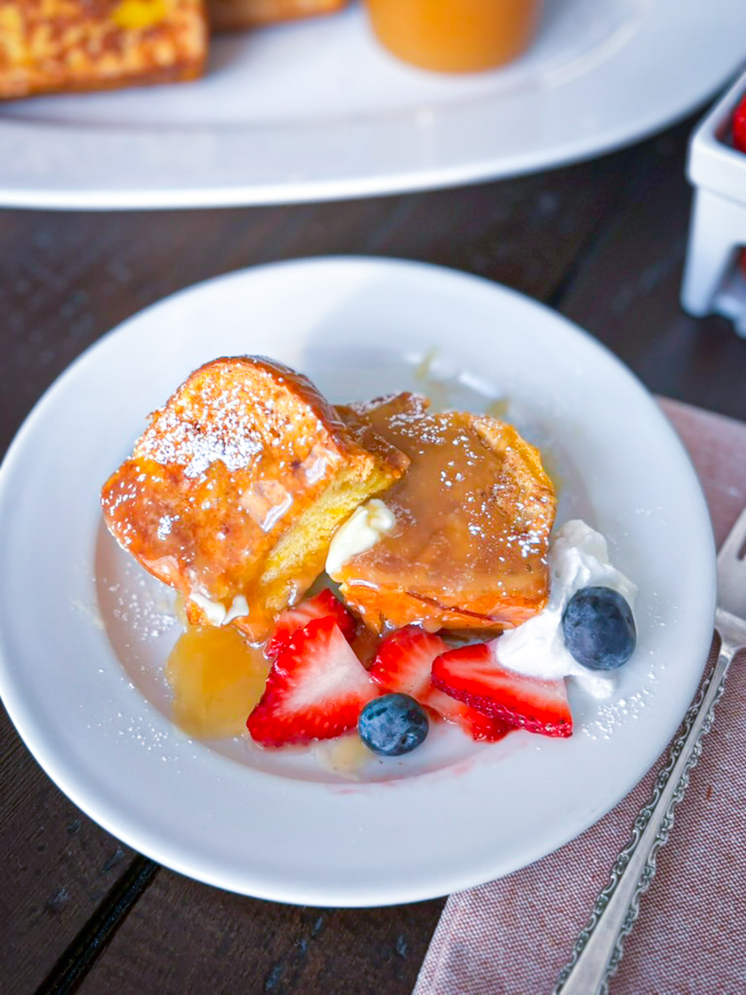 Thick slice of French toast drizzled with buttermilk syrup on a white plate with strawberries, blueberries and cream.