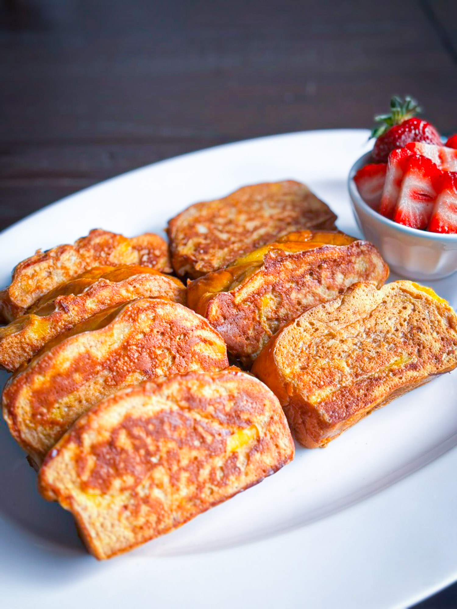 A plate of golden-brown French toast slices served with fresh strawberry slices in a small bowl.