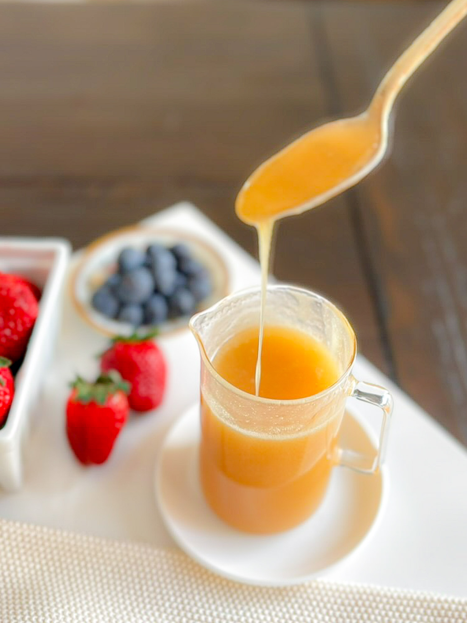 A glass pitcher filled with a golden liquid, with a spoon hovering above it, dripping the liquid. Surrounding the pitcher are fresh strawberries and a bowl of blueberries on a white surface.