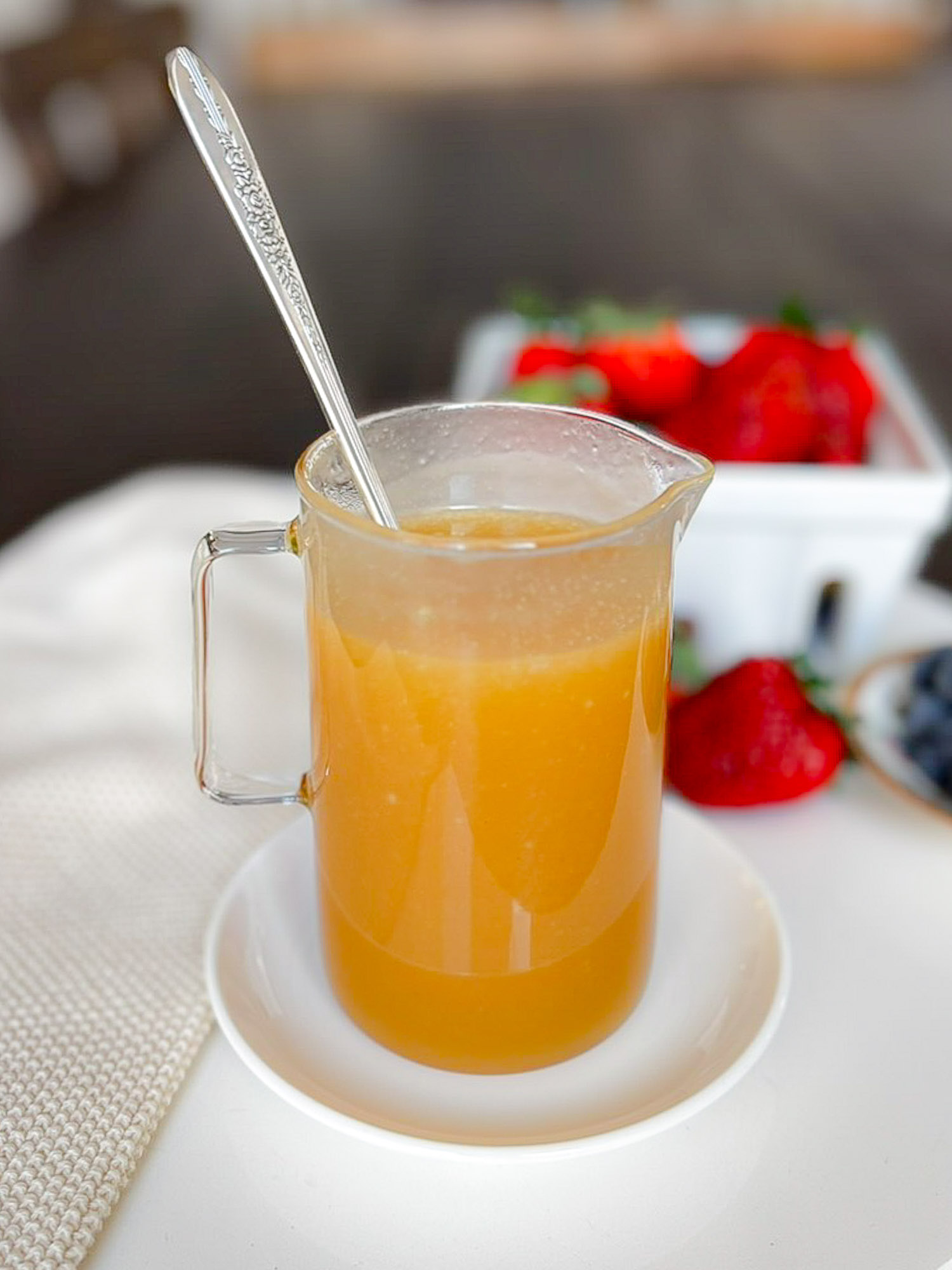 A glass pitcher filled with golden yellow liquid, accompanied by a silver spoon, and fresh strawberries and blueberries in the background.