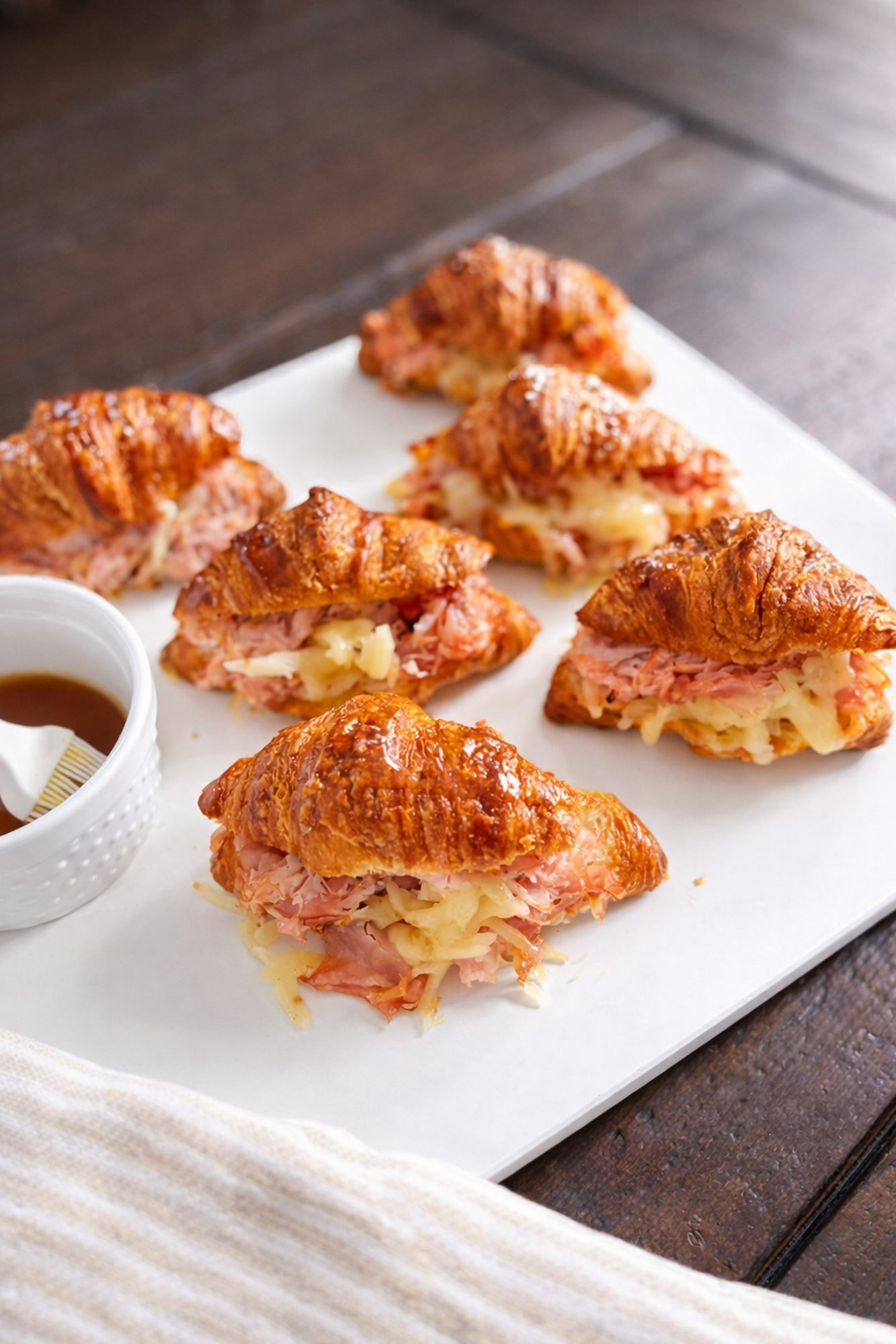 Baked Ham and Cheese Croissant Minis 4 Mini croissant sandwiches filled with ham and melted cheese on a white serving tray with a small bowl of sauce and brush
