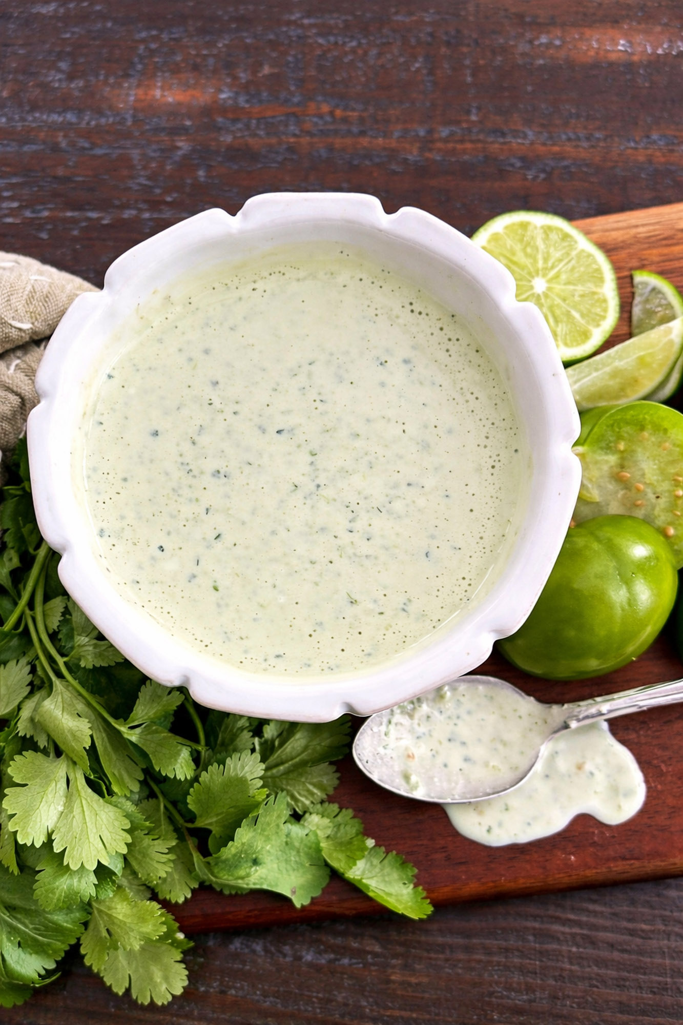 Jalapeno Tomatillo Ranch-20 White bowl filled with green creamy cilantro lime sauce on a wooden board with fresh cilantro and sliced limes