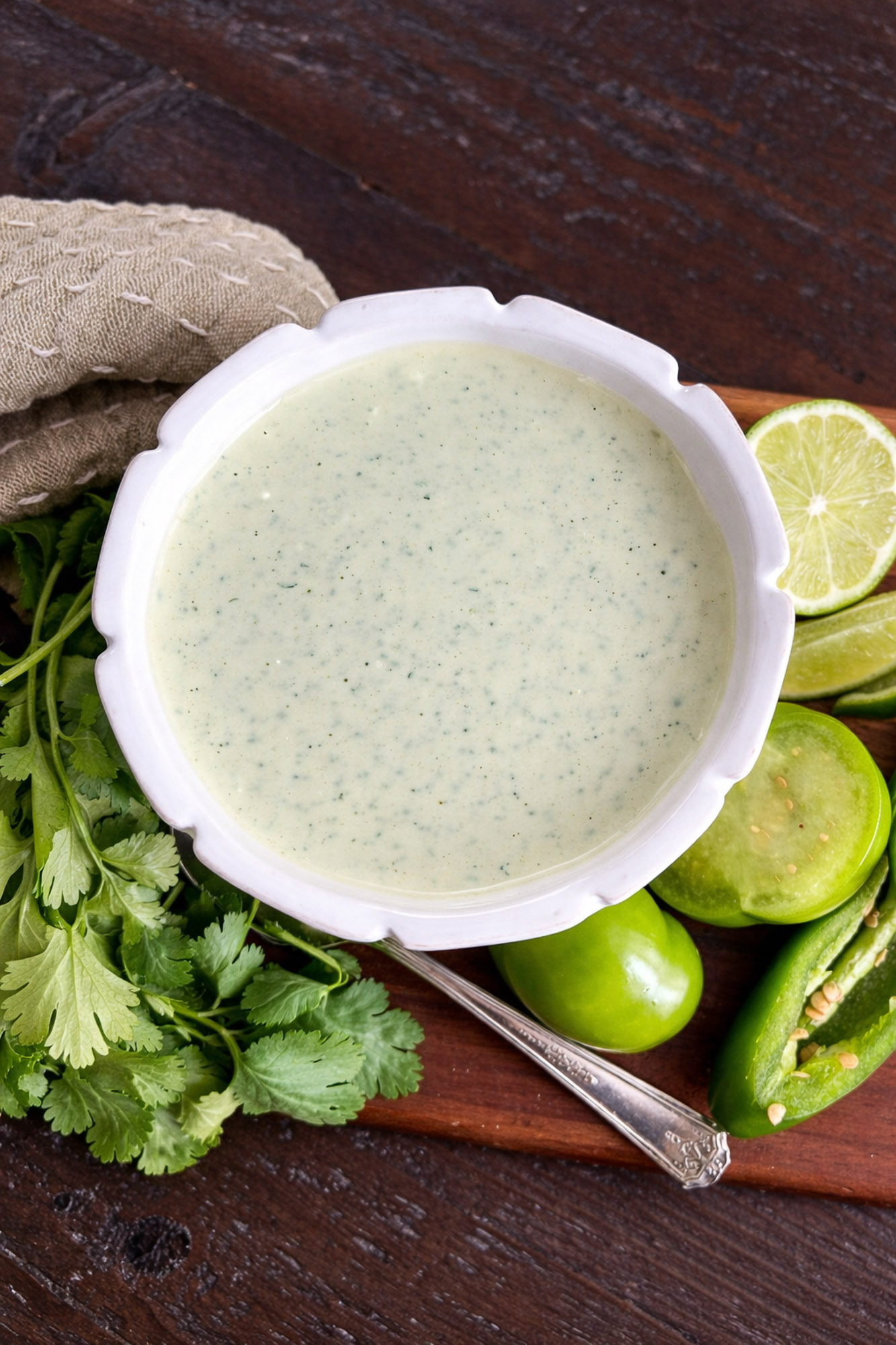 Jalapeno Tomatillo Ranch-22 White bowl of creamy green salsa verde sauce surrounded by fresh cilantro, lime slices, tomatillos, and jalapeño on a wooden surface