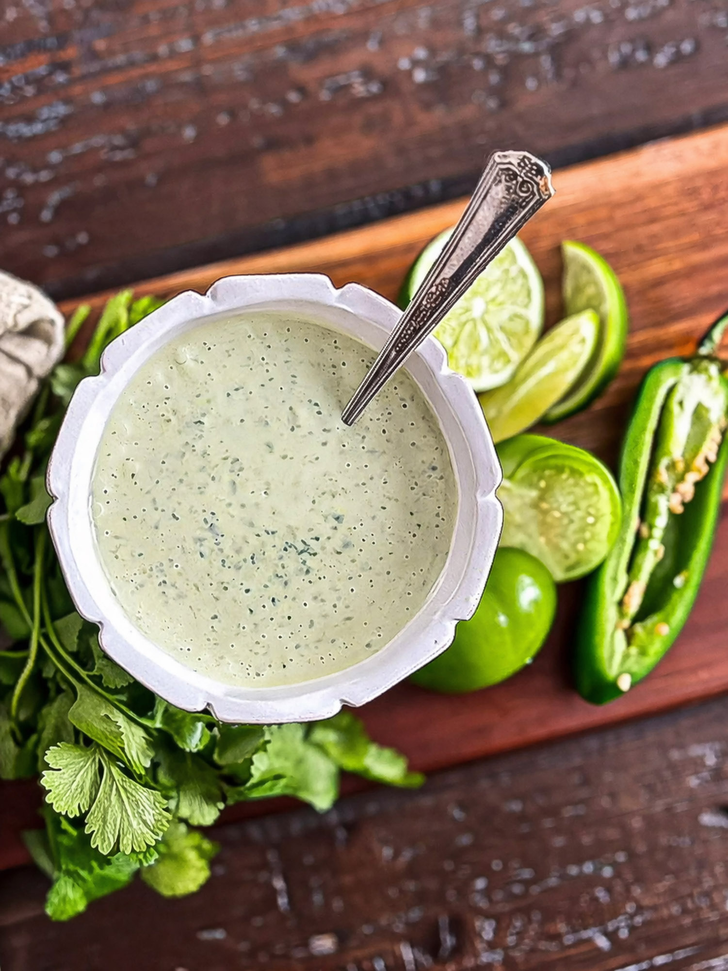 Jalapeno Tomatillo Ranch-23 White bowl of creamy cilantro lime sauce with spoon, fresh cilantro, lime slices, and sliced jalapeño on wooden surface