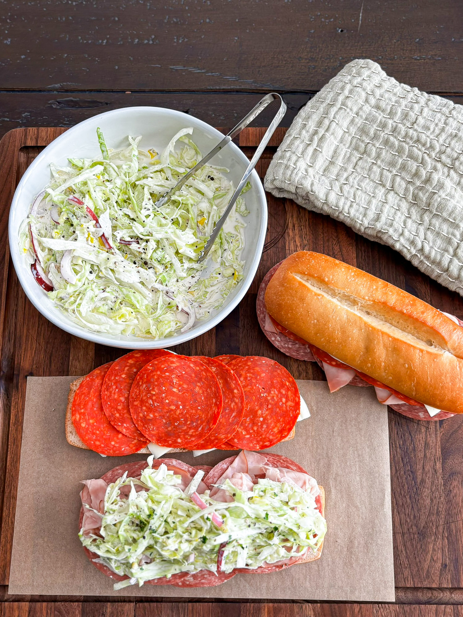 Italian Grinder Sandwich with Creamy Deli Slaw 1 Overhead view of Italian sandwiches, creamy salad, salami, and sliced bread on a wooden table
