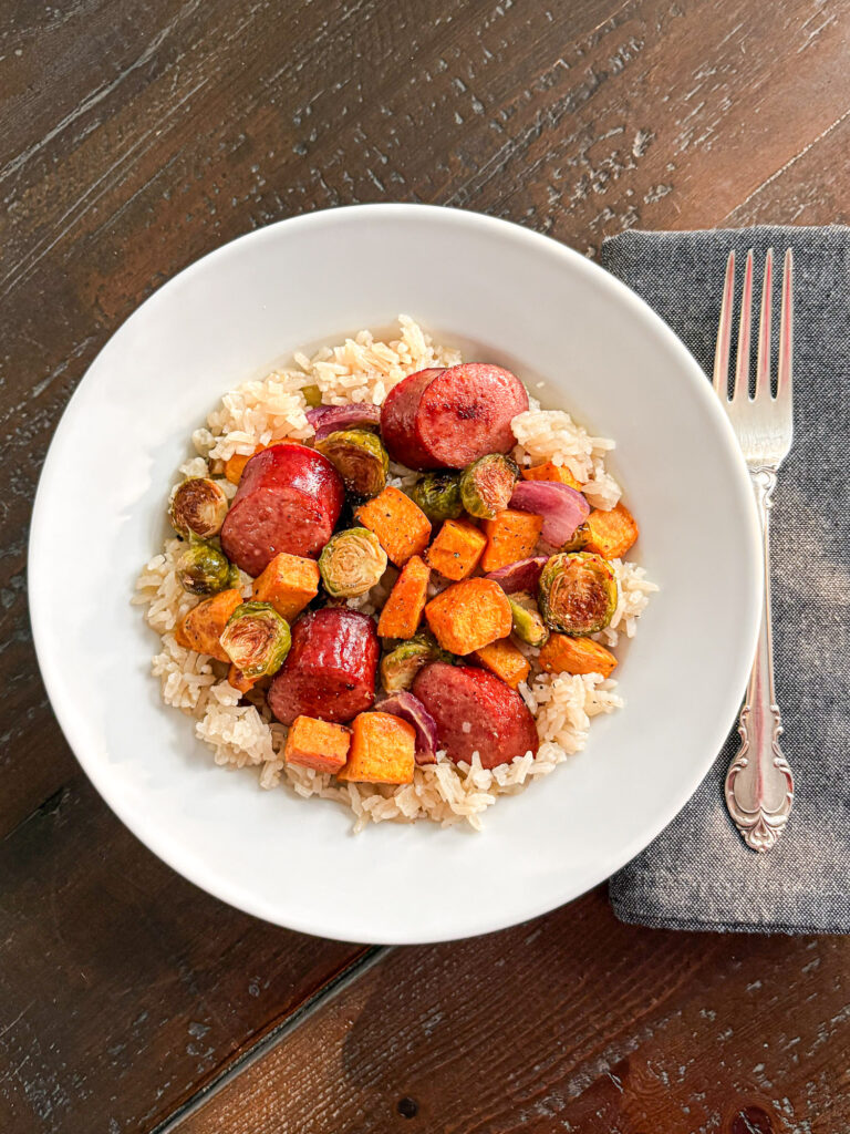 Overhead view of sliced smoked sausage, roasted sweet potatoes, Brussels sprouts, and red onion served over apple rice in a white bowl.